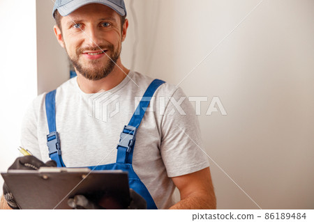 Portrait of electrical technician in uniform smiling at camera, writing the data collected on a residential electric panel Portrait of electrical technician in uniform smiling at camera, writing the data collected on a residential electric panel 86189484