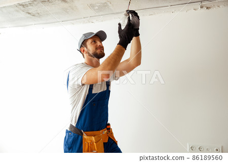 Focused electrician in uniform installing light fitting in new apartment, standing on a ladder Focused electrician in uniform installing light fitting in new apartment, standing on a ladder 86189560