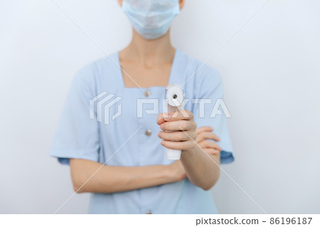 Temperature measurement gun in doctor hands. Close-up shot of doctor wearing protective surgical mask ready to use infrared isometric thermometer gun to check body temperature for virus symptoms. Temperature measurement gun in doctor hands. Close-up shot of doctor wearing protective surgical mask ready to use infrared isometric thermometer gun to check body temperature for virus symptoms. 86196187