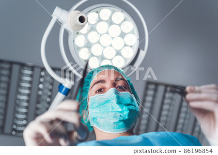 From below woman surgeon in medical uniform using professional tools while standing under bright From below woman surgeon in medical uniform using professional tools while standing under bright 86196854