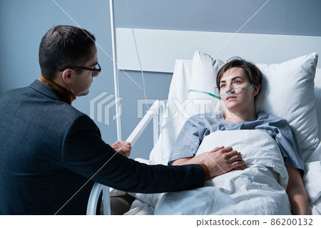 Rear view of young man supporting the sick young woman while she lying on bed at hospital ward 86200132