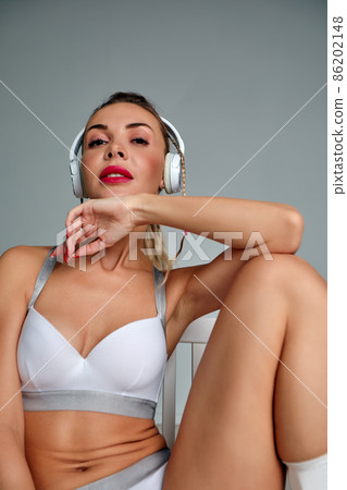 Beautiful girl in trendy white swimsuit having fun on gray background. Amazing blond-haired woman sitting on chair and listening music in headphones 86202148