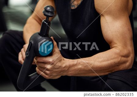 A young man is using and enjoying percussion massage gun to relax and relief the pain in body and muscles in the gym A young man is using and enjoying percussion massage gun to relax and relief the pain in body and muscles in the gym 86202335