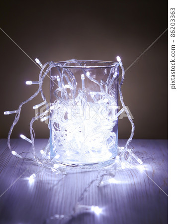 Christmas white garland lights from LED bulbs in glass jar on wooden table background. Front view 86203363