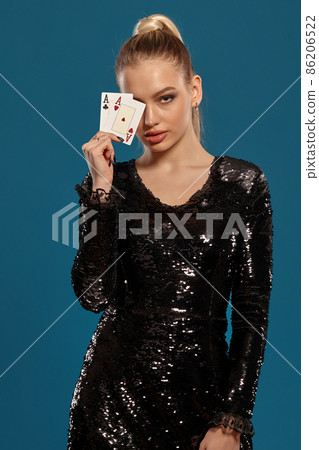 Blonde woman in black sequin dress showing two playing cards, posing against blue background. Gambling entertainment, poker, casino. Close-up 86206522