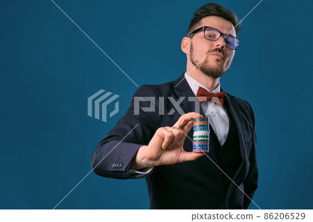 Man in black classic suit, red bow-tie, glases is showing some colored chips, posing on blue studio background. Gambling, poker, casino. Close-up. 86206529
