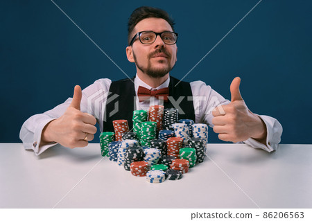 Player in glasses, black vest and shirt sitting at white table with stacks of chips on it, posing on blue background. Poker, casino. Close-up. Player in glasses, black vest and shirt sitting at white table with stacks of chips on it, posing on blue background. Poker, casino. Close-up. 86206563