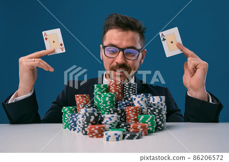 Man in glasses, black suit sitting at white table with stacks of chips, holding two playing cards, posing on blue background. Poker, casino. Close-up. Man in glasses, black suit sitting at white table with stacks of chips, holding two playing cards, posing on blue background. Poker, casino. Close-up. 86206572