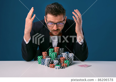 Man in glasses, black suit looking disappointed, sitting at white table with colored stacks of chips on it, posing on blue background. Poker, casino Man in glasses, black suit looking disappointed, sitting at white table with colored stacks of chips on it, posing on blue background. Poker, casino 86206578