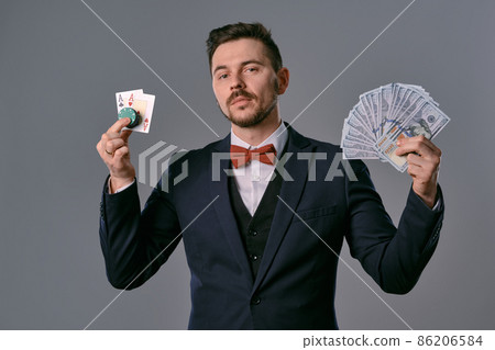 Man in black classic suit and red bow-tie showing two playing cards, chips and cash, posing on gray background. Gambling, poker, casino. Close-up. Man in black classic suit and red bow-tie showing two playing cards, chips and cash, posing on gray background. Gambling, poker, casino. Close-up. 86206584