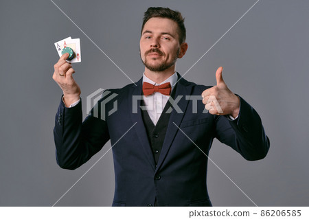 Man in black classic suit and red bow-tie showing two playing cards and chips, posing on gray studio background. Gambling, poker, casino. Close-up. Man in black classic suit and red bow-tie showing two playing cards and chips, posing on gray studio background. Gambling, poker, casino. Close-up. 86206585