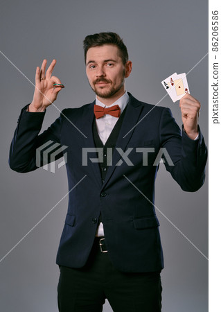 Man in black classic suit and red bow-tie showing two playing cards and chips, posing on gray studio background. Gambling, poker, casino. Close-up. Man in black classic suit and red bow-tie showing two playing cards and chips, posing on gray studio background. Gambling, poker, casino. Close-up. 86206586