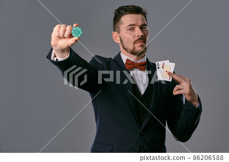 Man in black classic suit and red bow-tie showing two playing cards and chip, posing on gray studio background. Gambling, poker, casino. Close-up. Man in black classic suit and red bow-tie showing two playing cards and chip, posing on gray studio background. Gambling, poker, casino. Close-up. 86206588