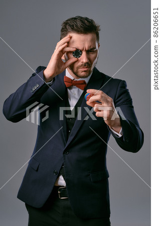 Man in black classic suit and red bow-tie showing some colored chips, posing against gray studio background. Gambling, poker, casino. Close-up. Man in black classic suit and red bow-tie showing some colored chips, posing against gray studio background. Gambling, poker, casino. Close-up. 86206651