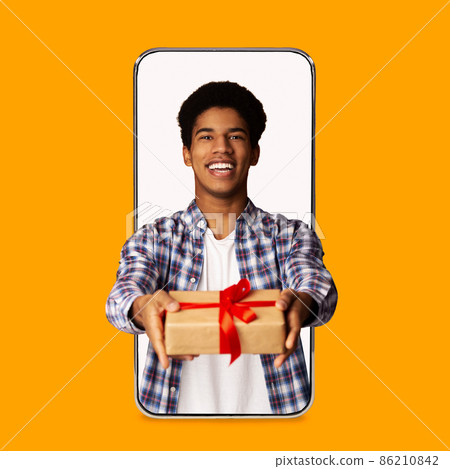 Happy young attractive black guy gives gift in box on blank screen of big phone isolated on orange background 86210842
