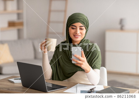 Arab woman in hijab using laptop and smartphone for distance job, drinking coffee while communicating online from home 86211068