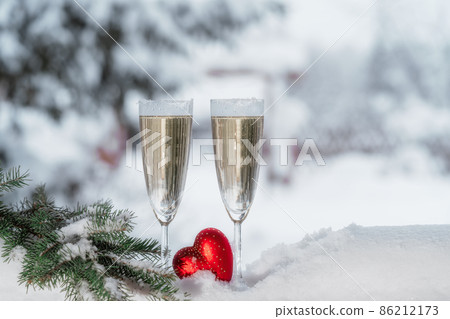 Closeup of two champagne flutes cooling in snow and red heart on winter nature background. Valentine's day celebration concept. Copy space. Closeup of two champagne flutes cooling in snow and red heart on winter nature background. Valentine's day celebration concept. Copy space. 86212173