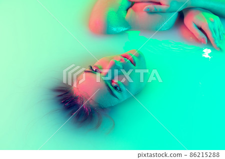 Top view of female face, young girl in the milk bath with soft glowing in blue-green neon light. Beauty, fashion, style, skincare concept 86215288