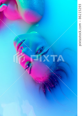 Top view of female face, young girl in the milk bath with soft glowing in blue-pink neon light. Beauty, fashion, style, skincare concept Top view of female face, young girl in the milk bath with soft glowing in blue-pink neon light. Beauty, fashion, style, skincare concept 86215293