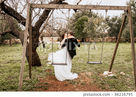 Happy stylish smiling couple walking in Tuscany, Italy on their wedding day. NEWLYWEDS WITH THE SWING IN THE PARK. The bride and groom walk down the street by the hands. A stylish young couple walks 86216795