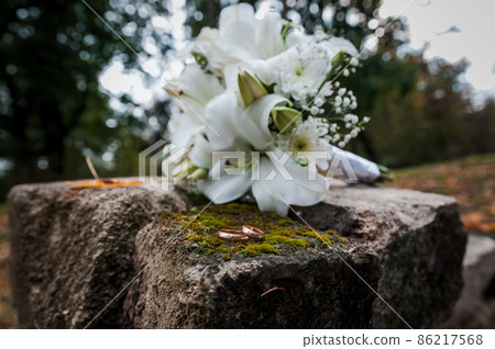 Floral arrangement with white flowers, wedding bouquet with a ribbon and two gold rings, lying on a stone. Wedding decor, artwork, florist 86217568