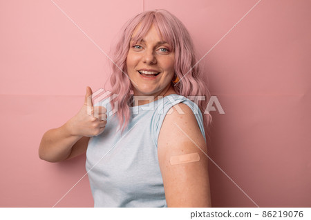Coronavirus vaccinated woman showing thumb sign with bandage after receiving covid 19 vaccine isolated over pink wall background. 86219076