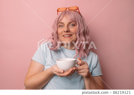 Attractive beautiful female woman hand holds hot cup coffee drink and teapot tea look at camera enjoying sunny morning isolated over pink wall background. 86219121