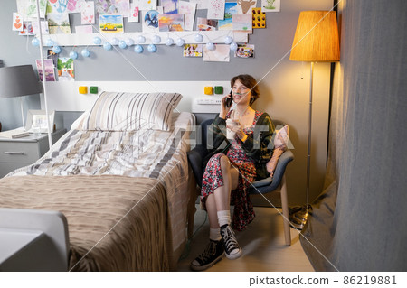 Happy teenage girl talking on mobile phone and having drink while relaxing in armchair Happy teenage girl talking on mobile phone and having drink while relaxing in armchair 86219881