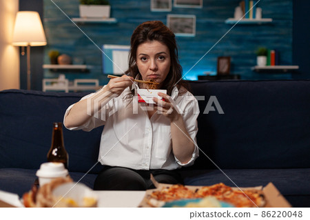 Tired woman holding chopsticks and chinese takeaway noodle box sitting on couch 86220048