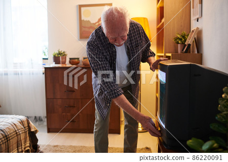 Senior man bending by tv set while switching it on or off Senior man bending by tv set while switching it on or off 86220091