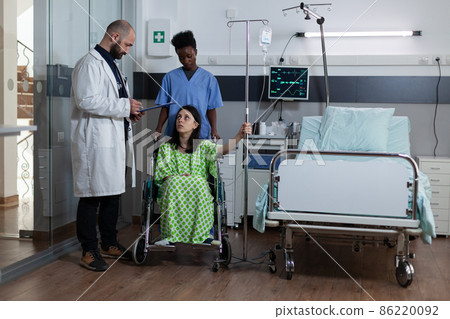 Patient with illness sitting in wheelchair holding drip stand in modern hospital ward 86220092