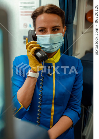 Flight attendant in medical mask talking on telephone in airplane Flight attendant in medical mask talking on telephone in airplane 86220126