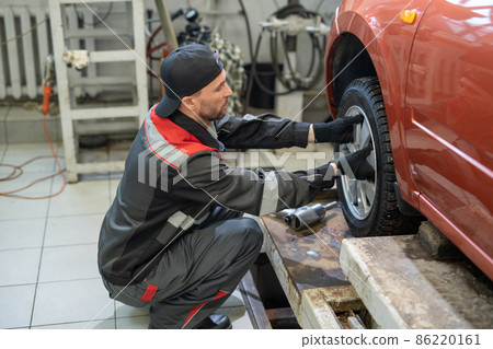 Young repairman in workwear putting new wheel in car Young repairman in workwear putting new wheel in car 86220161
