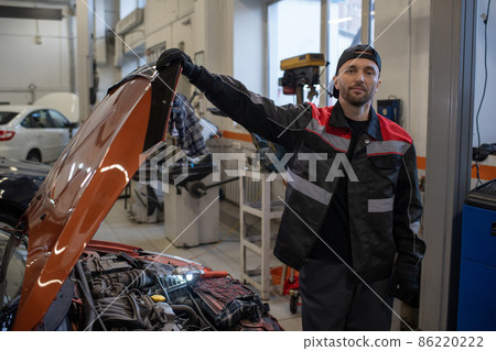 Young male mechanic in workwear opening cover of car hood 86220222