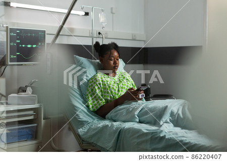 African american sick patient lying in bed holding modern phone browsing on internet 86220407