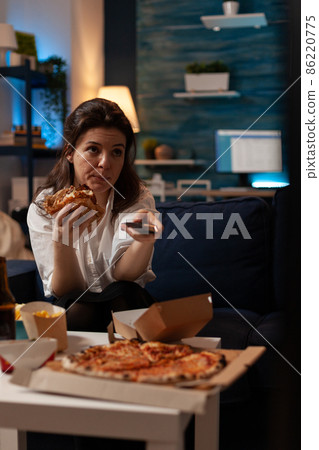 Woman sitting on sofa eating tasty delicious burger holding remote control switching channels on television 86220775