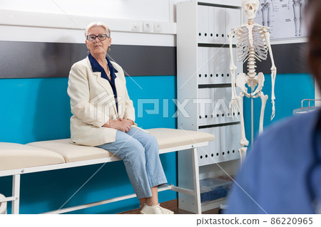 Portrait of sick sick pensioner woman patient standing on bed in hospital office looking into camera 86220965