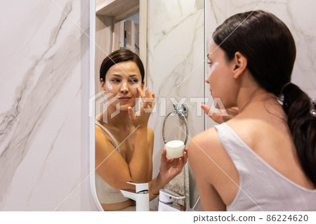Happy domestic woman applying anti aging cream from jar on face looking at mirror in bathroom Happy domestic woman applying anti aging cream from jar on face looking at mirror in bathroom 86224620