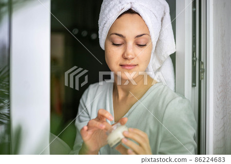 Happy domestic woman applying anti aging cream from jar on face looking at mirror Happy domestic woman applying anti aging cream from jar on face looking at mirror 86224683