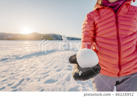 Winter outdoor active lifestyle concept. Girl holding snowball on snowy day with snowman in background on frozen lake at beautiful winter sunset Winter outdoor active lifestyle concept. Girl holding snowball on snowy day with snowman in background on frozen lake at beautiful winter sunset 86228952