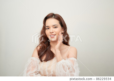 Tender asian woman wearing natural makeup, white blouse, touching her facial skin and gazing at camera Tender asian woman wearing natural makeup, white blouse, touching her facial skin and gazing at camera 86229334