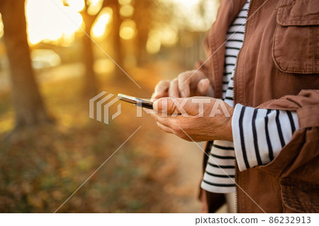 woman hand holding mobile cellphone, reading news or watching online having video call using mobile application. Beautiful autumn nature background woman hand holding mobile cellphone, reading news or watching online having video call using mobile application. Beautiful autumn nature background 86232913