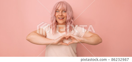 Charming young caucasian pink hair woman feels happy and romantic shapes heart gesture poses against pink background. Charming young caucasian pink hair woman feels happy and romantic shapes heart gesture poses against pink background. 86232987