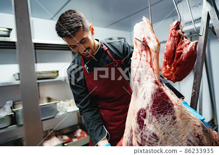 Male butcher standing by meat hanging on hook in meat shop 86233935