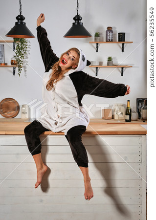 Blonde female in plush pajama in form of cartoon character panda is having fun at bachelorette party. Yawning, sitting on kitchen table. Close-up. Blonde female in plush pajama in form of cartoon character panda is having fun at bachelorette party. Yawning, sitting on kitchen table. Close-up. 86235549