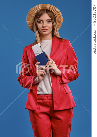 Blonde woman in straw hat, white blouse, red pantsuit. She is holding passport and ticket, posing on blue background. Travelling concept. Close-up Blonde woman in straw hat, white blouse, red pantsuit. She is holding passport and ticket, posing on blue background. Travelling concept. Close-up 86235707