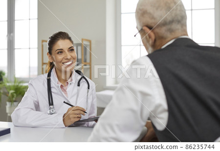 Friendly young female doctor in hospital office consults senior male patient and makes entries in his medical card. Smiling nurse sitting at table talking to mature man sitting with his back to camera Friendly young female doctor in hospital office consults senior male patient and makes entries in his medical card. Smiling nurse sitting at table talking to mature man sitting with his back to camera 86235744