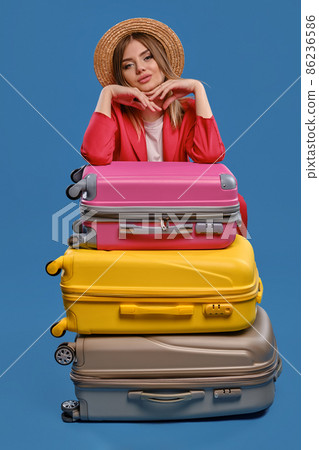 Blonde lady in straw hat, white blouse, red jacket. Sitting leaning on colorful suitcases, posing on blue background. Travelling concept. Close-up Blonde lady in straw hat, white blouse, red jacket. Sitting leaning on colorful suitcases, posing on blue background. Travelling concept. Close-up 86236586