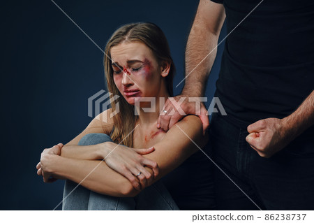 Strong man put his hand on female shoulder, showing fist. Victim with bruises on face sitting nearby. Blue background. Domestic violence. Close-up. 86238737