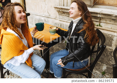 Cheerful female best friend share multimedia files on smartphones spending free time together,smiling hipster girls drinking coffee, sitting at terrace cafe. 86239571